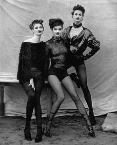 three women are posing for the camera in front of a white sheeted backdrop, wearing short skirts and high heels