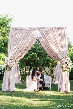 a bride and groom sitting on a white couch under an outdoor wedding ceremony set up