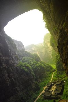 the inside of a cave with green grass and trees on both sides, looking down at an area that has houses built into it