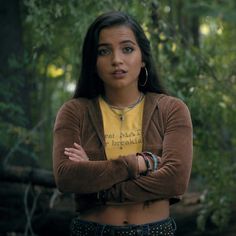 a woman with her arms crossed standing in front of some trees and looking at the camera