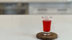 a glass filled with red liquid sitting on top of a white counter next to a wooden coaster