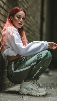 a woman with red hair is leaning against a brick wall wearing green pants and white sneakers