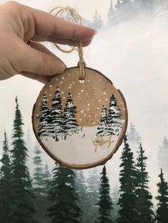 a hand holding a wooden ornament with trees on it and snow falling off the tops