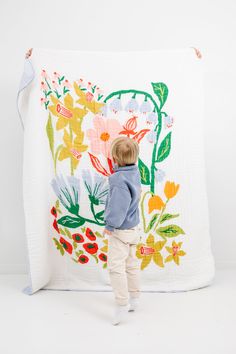 a little boy standing in front of a flowered wall hanging on a white quilt