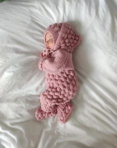 a baby in a pink knitted outfit laying on top of a white bed next to pillows