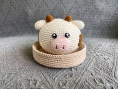 a small crocheted cow head sitting in a bowl on a gray surface, with eyes wide open