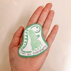 a hand holding a green and white sticker with a dinosaur on it