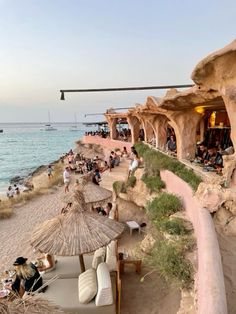 many people are sitting on the beach by the water and some have straw umbrellas