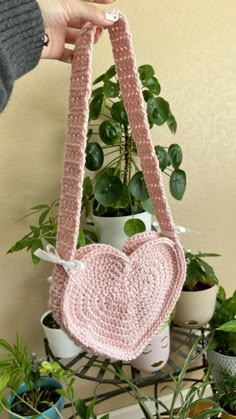 a hand holding a pink crocheted heart shaped purse next to potted plants