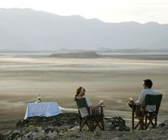 two people sitting at a table on top of a rocky hill overlooking the ocean and mountains