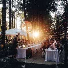 a group of people sitting at tables in the woods with lights strung from trees behind them