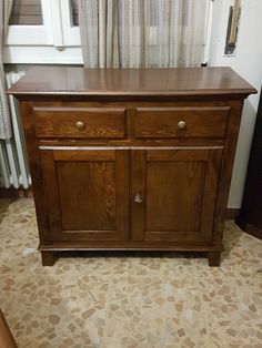 a wooden cabinet with two doors and one drawer on the bottom, in front of a curtained window