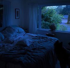 a bed in a dark room with blue light coming through the window and trees outside