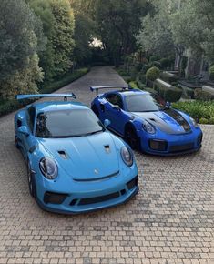 two blue sports cars parked next to each other on a brick road in front of trees