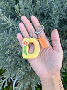 a hand holding a keychain shaped like the letter d with an orange flower on it