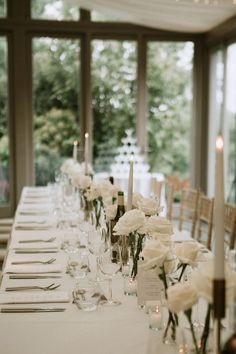 a long table is set with white flowers and candles for an elegant wedding reception in front of large windows