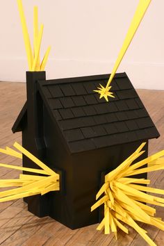 a black house with yellow sticks sticking out of it's roof and windows, on a wooden floor