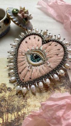 a pink brooch with an eye on it and pearls around the eyes is sitting next to a cup