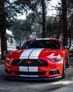 the front end of a red and white mustang parked in a parking lot next to trees