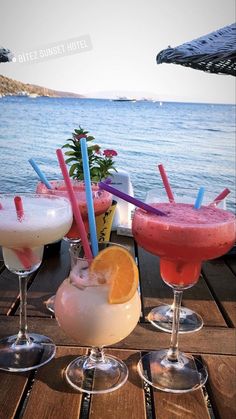 three different colored drinks sitting on top of a wooden table next to the ocean,