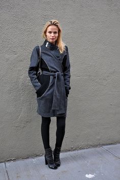 a woman standing in front of a gray wall wearing black boots and a grey coat