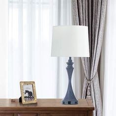 a table with a lamp on it next to a curtained window and a wooden dresser