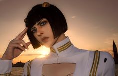 a woman with short black hair wearing a white shirt and gold trims standing in front of the ocean at sunset