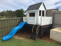 a small house with a blue slide in the yard next to a wooden fence and green grass