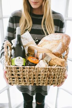 a woman holding a wicker basket filled with bread, wine and other food items