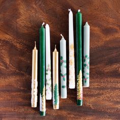 five candles are lined up on a wooden table with holly and pine trees painted on them