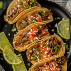 four shredded beef tacos on a black plate with limes and cilantro