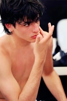 a shirtless young man sitting at a table with his hand on his chin and looking off to the side