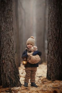 a little boy standing in the woods with a teddy bear on his back and holding a lantern