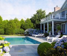 a pool surrounded by bushes and flowers next to a large white house with columns in the background