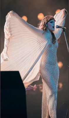 a woman in white dress standing on stage with her arms spread out to the side