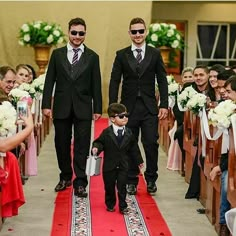 two men in suits and sunglasses are walking down the aisle with their young son, who is carrying a suitcase