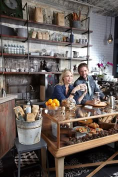 a man and woman sitting at a table in a kitchen with lots of food on it