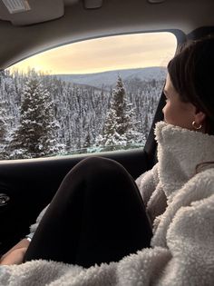 a woman sitting in the passenger seat of a car looking out at snow covered trees