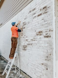 a man in an orange shirt is on a ladder painting a brick wall with white paint