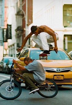 two men riding bikes in the street with cars behind them and one man doing tricks on his bike