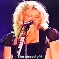 a woman singing into a microphone while wearing a black dress and holding a guitar in her hand