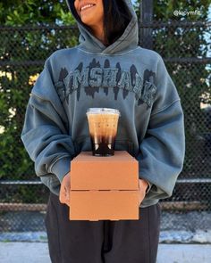 a woman holding a cardboard box with a drink in it's hand and wearing a hoodie