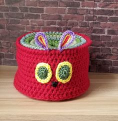 a red crocheted hat with eyes and ears on the top, sitting on a wooden table