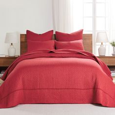 a bed with red comforter and pillows in a white room next to a window