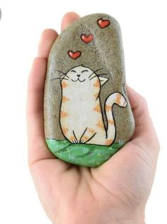 a hand holding up a rock with a cat painted on it's face and hearts flying above