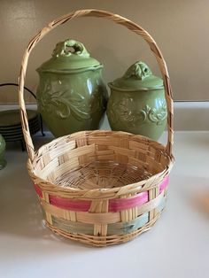 a basket and two jars sitting on a counter next to each other, one has a pink ribbon around the handle