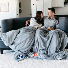 a man and woman sitting on a couch wrapped up in blankets smiling at each other