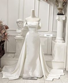 a white wedding dress on display in a room with statues and vases behind it