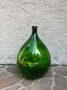 a green glass vase sitting on top of a stone floor next to a white wall