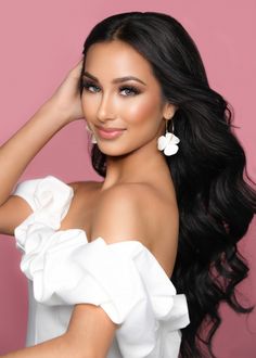 a woman with long black hair wearing a white dress and flower earrings on her head